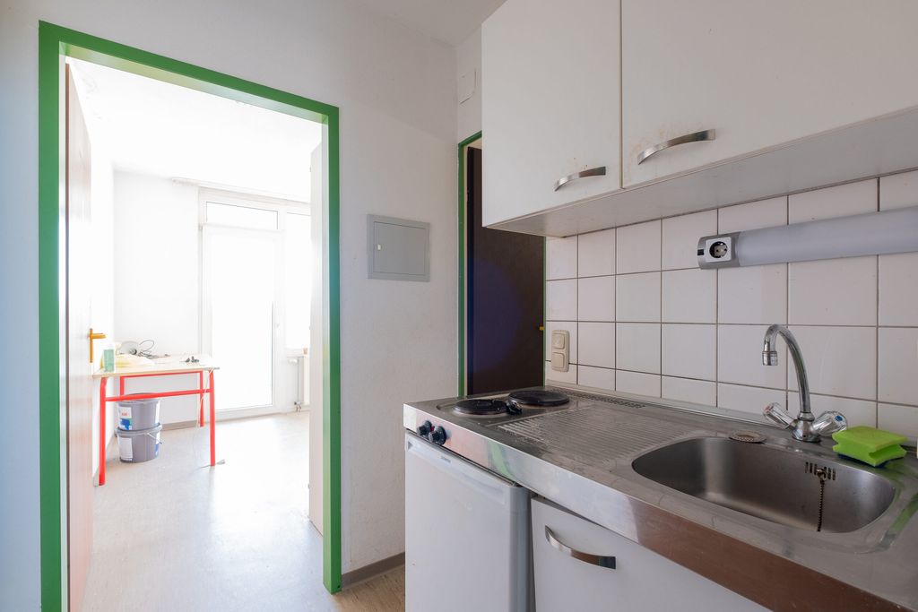 Kleine Küchenzeile mit Edelstahlspüle, Herdplatten und weißen Schränken an der Wand, Blick in einen hellen Raum mit Tisch und Töpfen im Hintergrund.