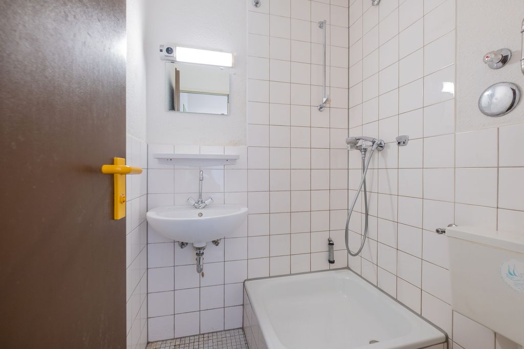 Kleines Badezimmer mit weiß gefliester Dusche, weißem Waschbecken unter einem kleinen Spiegel und einer halboffenen braunen Tür mit gelbem Türgriff.
