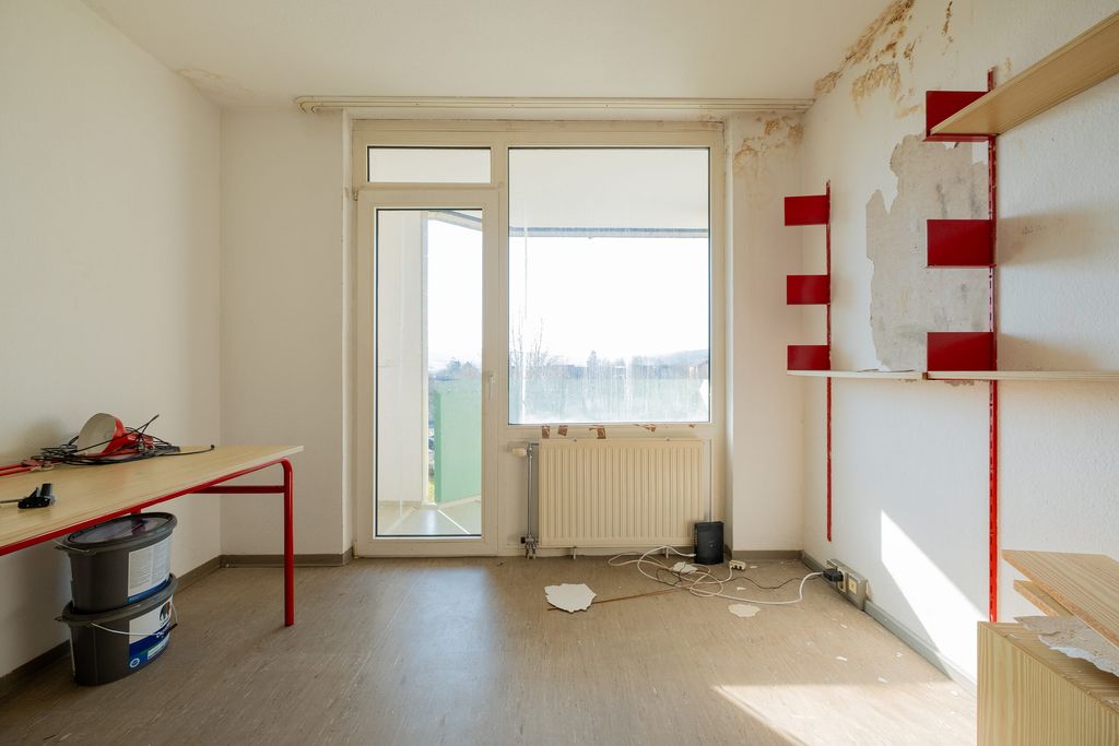 Leerer Raum mit einem Tisch an der linken Wand, beschädigter weißer Wand mit roten Regalen an der rechten Seite und einem großen Fenster mit Tür zum Balkon im Zentrum des Bildes.