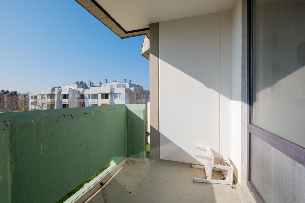 Ein leerer Balkon mit grün gestrahltem Geländer und einem umgestürzten weißen Stuhl unter blauem Himmel, mit Wohngebäuden im Hintergrund.