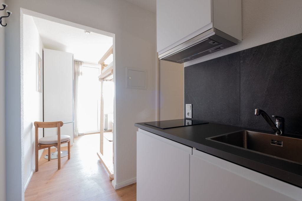 Moderne, minimalistische Küchenzeile mit schwarzer Arbeitsplatte, Spüle und Dunstabzug, angrenzend an einen hellen Flur mit Holzstuhl und großem Fenster.