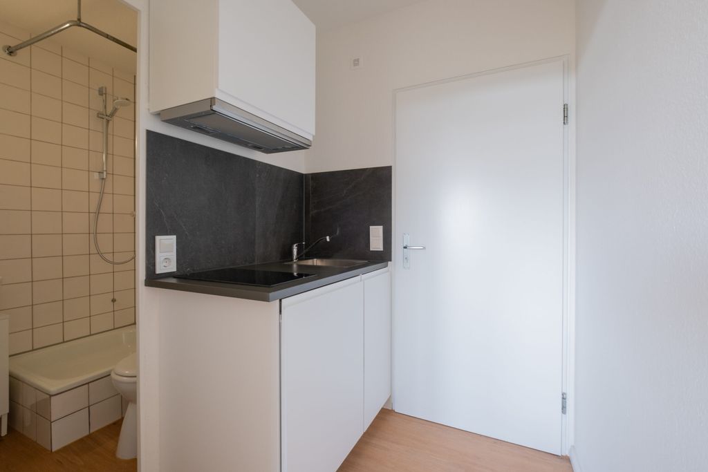 Kleine moderne Küchenzeile mit weißem Schrank, schwarzer Arbeitsplatte und Spüle in einem weißen Raum neben einer geschlossenen weißen Tür und einer Badezimmerdusche.