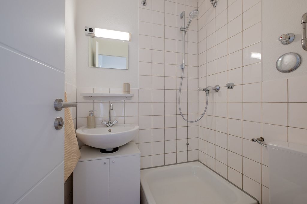 Kleines Badezimmer mit weißer Dusche, Waschbecken auf einem Unterschrank, klein gespiegelt und einem Handtuch an der Tür.