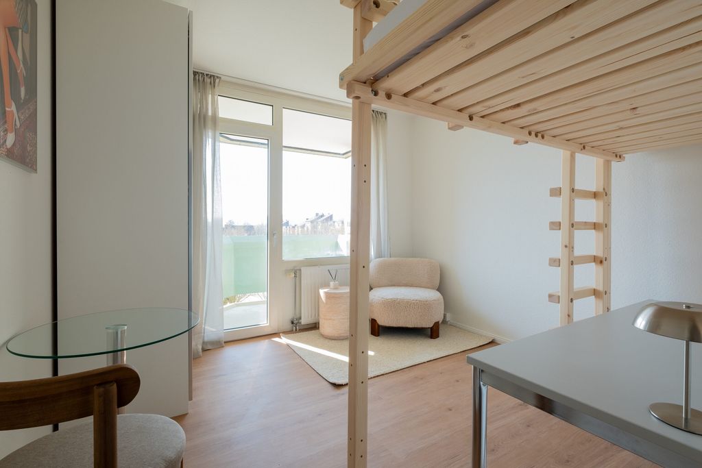 Heller, minimalistischer Raum mit Holz-Hochbett, beigem Sessel, Glasschreibtisch und Zugang zu einem Balkon mit Fensterfront.