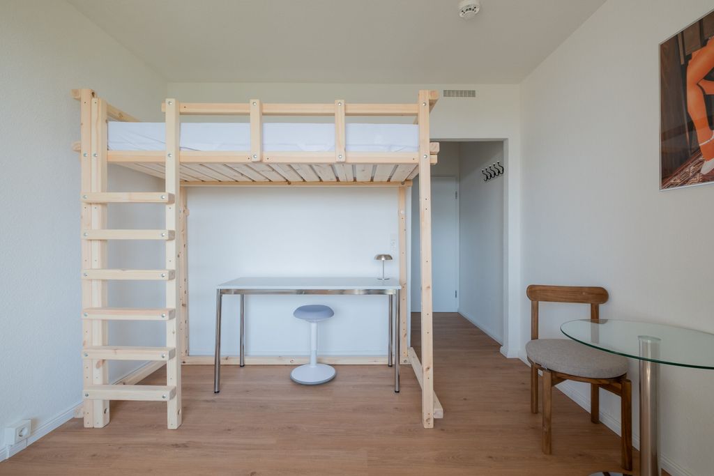 Ein schlicht eingerichtetes Zimmer mit einem Hochbett aus hellem Holz, darunter ein Tisch mit einem blauen Hocker und einer kleinen Lampe, sowie ein Holzhocker neben einem Glastisch an der rechten Wand.