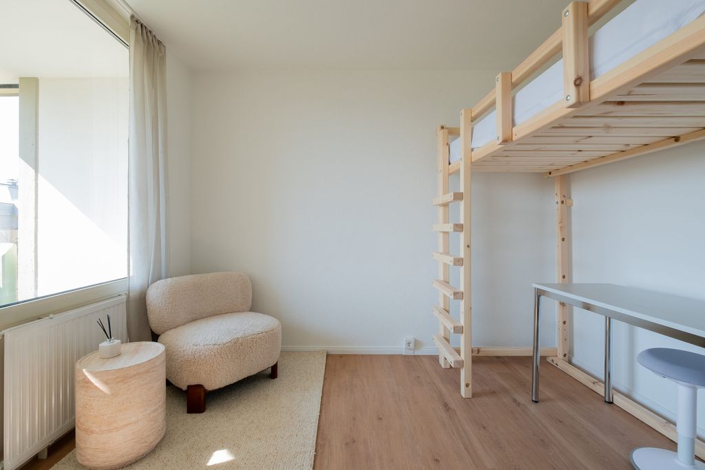 Ein minimalistischer Raum mit einem kleinen beigen Sessel neben einem runden Beistelltisch vor einem großen Fenster und einem Hochbett aus hellem Holz mit einem weißen Schreibtisch und Hocker darunter.