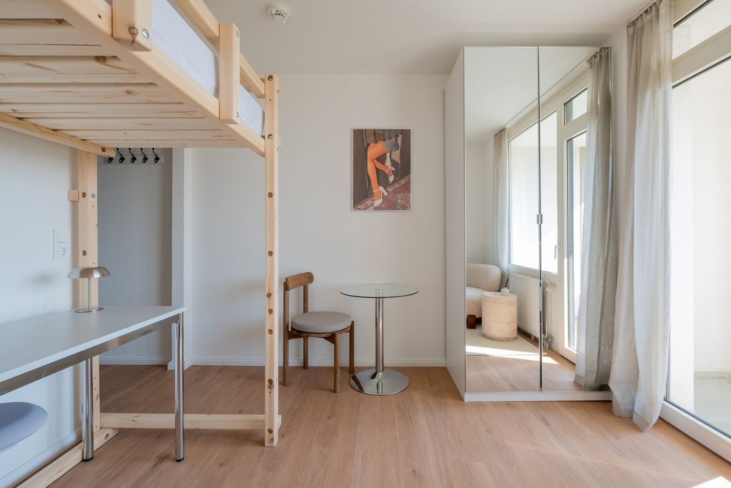 Helles Schlafzimmer mit Hochbett aus hellem Holz, einem kleinen runden Glastisch, einem Stuhl und einem Kleiderschrank mit Spiegeltüren neben einem großen Fenster mit Vorhängen.