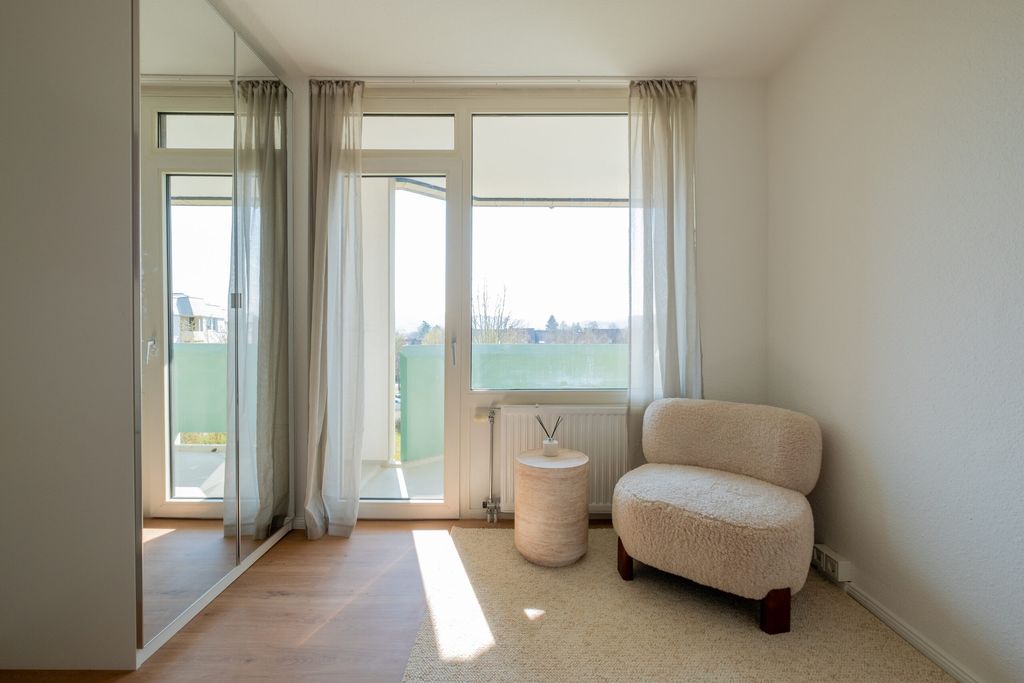 Heller, minimalistischer Wohnraum mit großen Fenstern und einer Tür zum Balkon, einem cremefarbenen Sessel und einem kleinen runden Beistelltisch auf einem hellen Teppich.