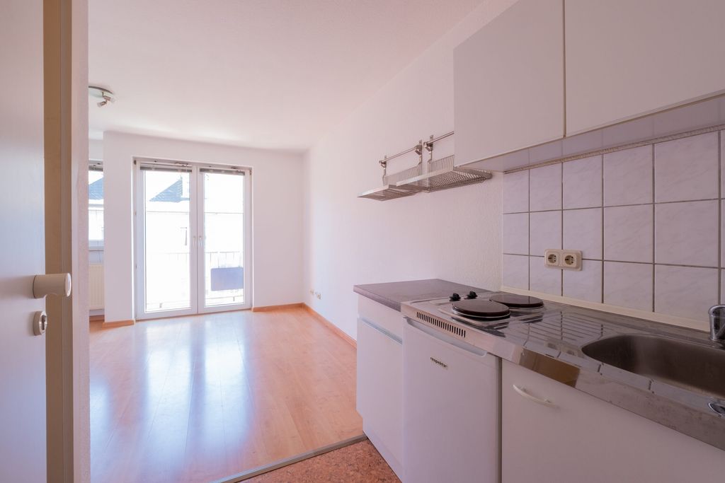 Leere, helle Wohnung mit Küchenzeile, Elektroherd, Spüle und großer Fensterfront mit Balkonzugang.