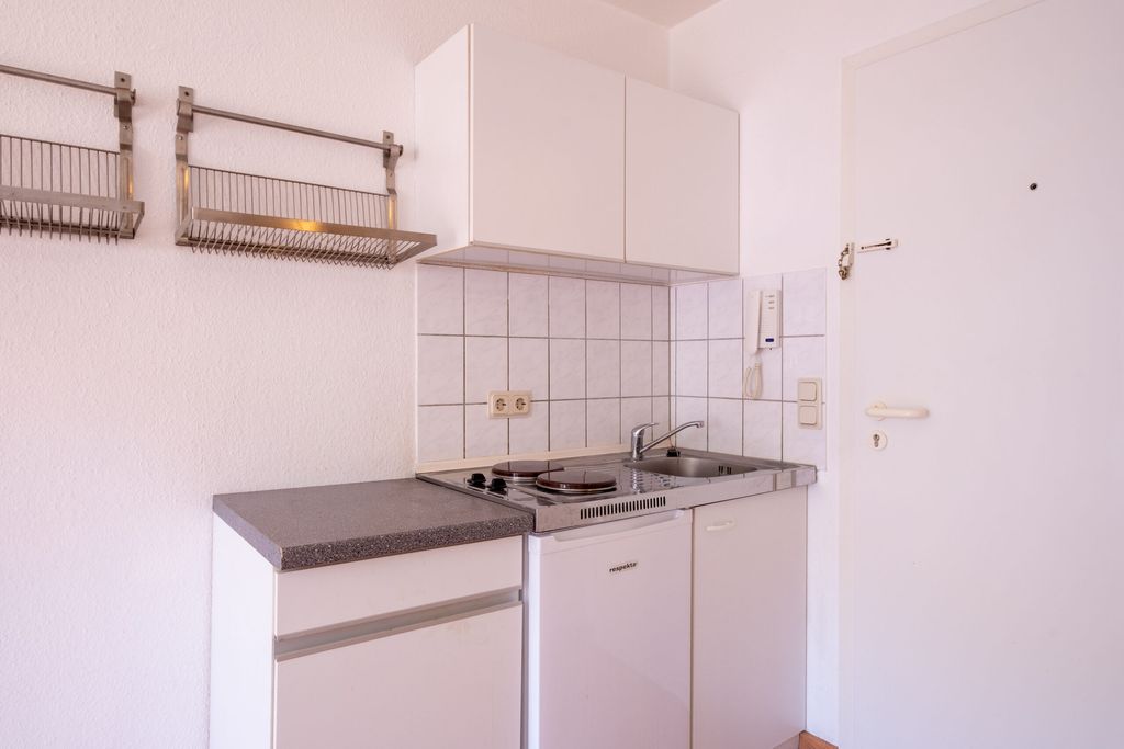 Kleine weiße Küchenzeile mit Herd, Spüle und Kühlschrank in einem einfachen Raum mit weißer Tür und Fliesenspiegel.