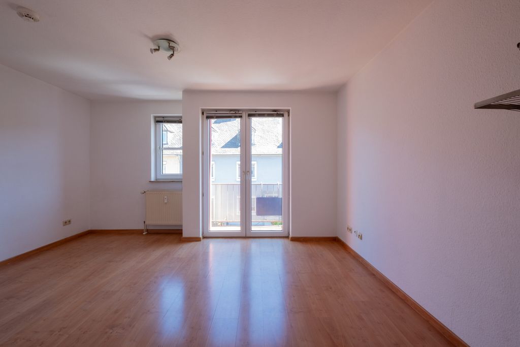Leerer Raum mit hellen Wänden, Holzboden, einer kleinen Fensteröffnung und einer gläsernen Doppeltür mit Blick auf ein Nachbargebäude.