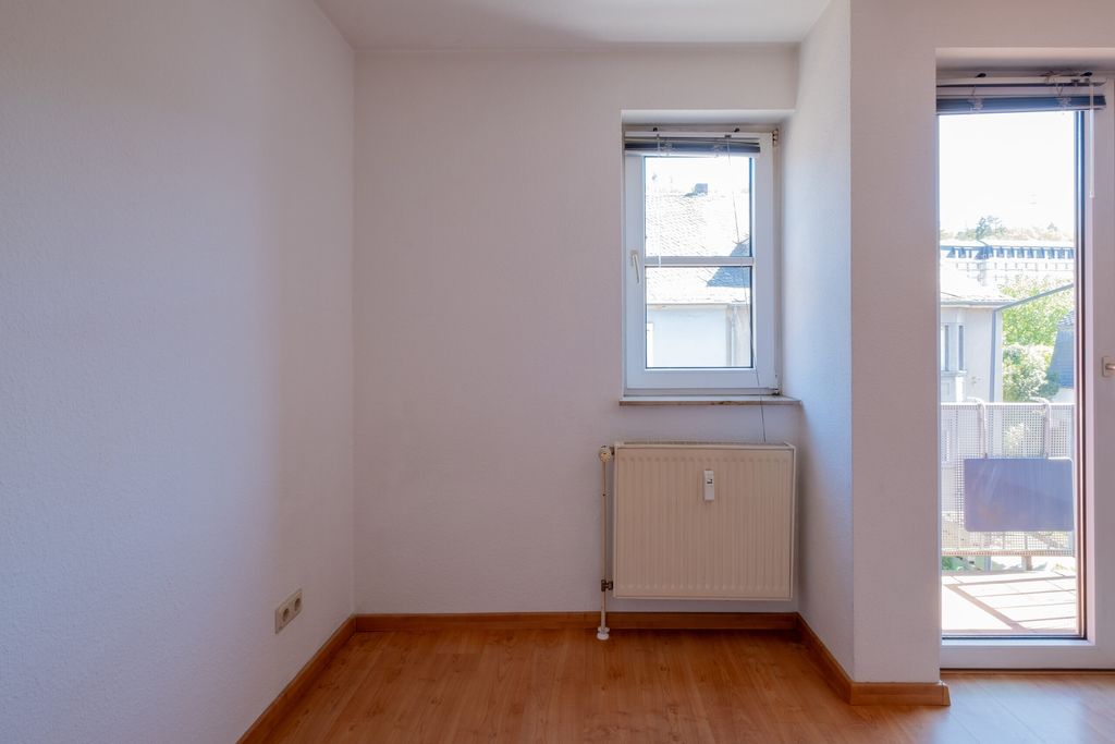 Leerer Raum mit Holzfußboden, weiß gestrichenen Wänden, einem Heizkörper unter einem kleinen Fenster und einer Terrassentür mit Blick auf eine Außenfläche.