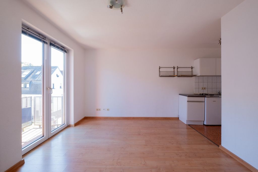 Leerer Raum mit Holzboden und großen Fenstertüren, die auf einen Balkon mit Blick auf angrenzende Häuser führen, sowie eine kleine Küchenzeile mit weißen Schränken und Fliesenspiegel.