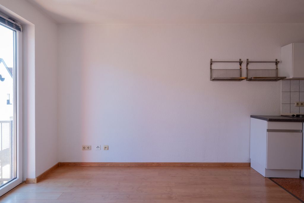 Leerer Raum mit Holzboden, weißen Wänden und Kühlschrank neben einer Küchenzeile mit angeordnetem Geschirrkorb, große Glastür links im Bild.