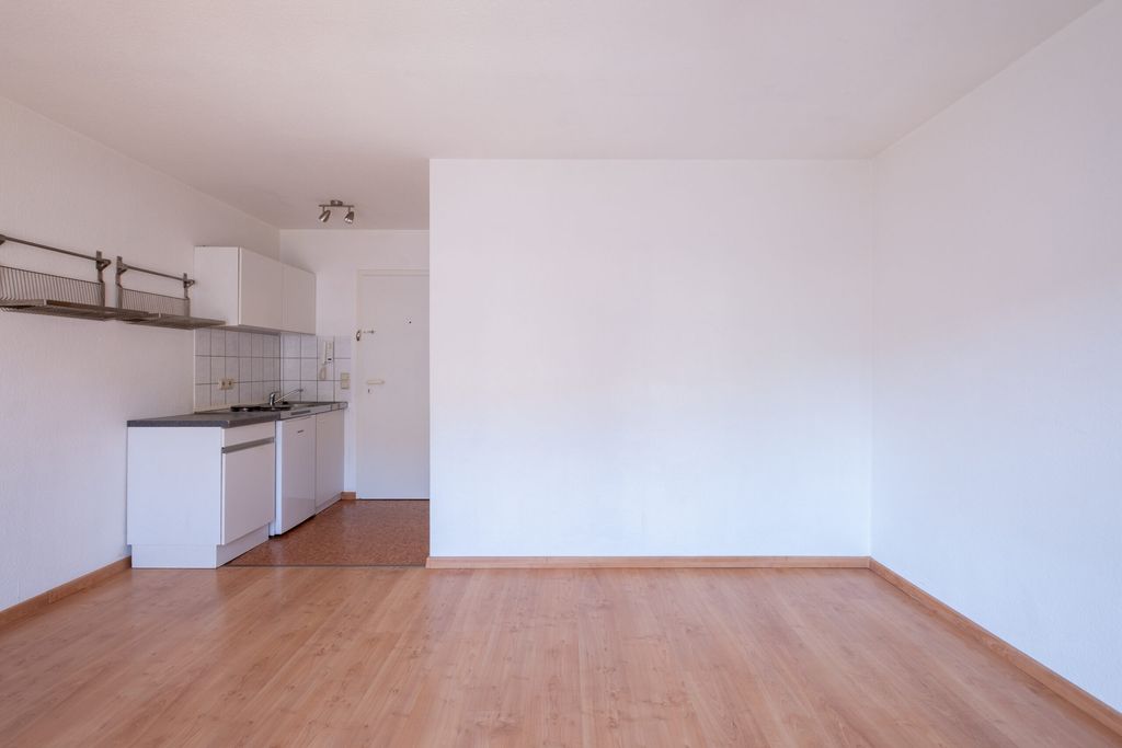 Leerer Raum mit hellen Holzboden und einer kleinen Küche mit weißen Schränken und Fliesen an der Wand links, daneben eine weiße Tür.