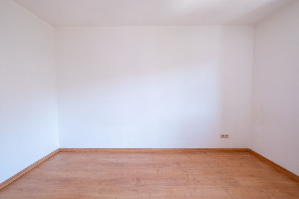 Leerer Raum mit weißen Wänden und hellem Holzfußboden, eine Steckdose an der rechten Wand sichtbar.