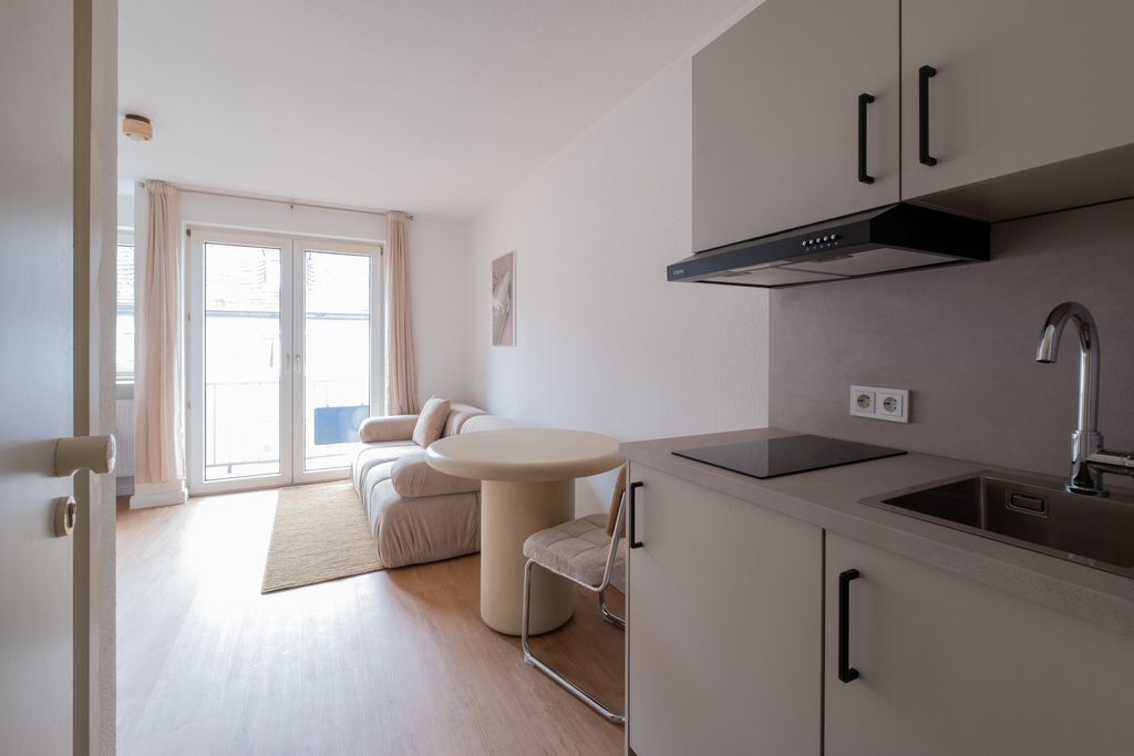 Helles Wohnzimmer mit großen Fenstertüren, einem beigen Sofa, einem runden weißen Tisch und einer modernen Küchenzeile mit Herd, Abzugshaube und Spüle.