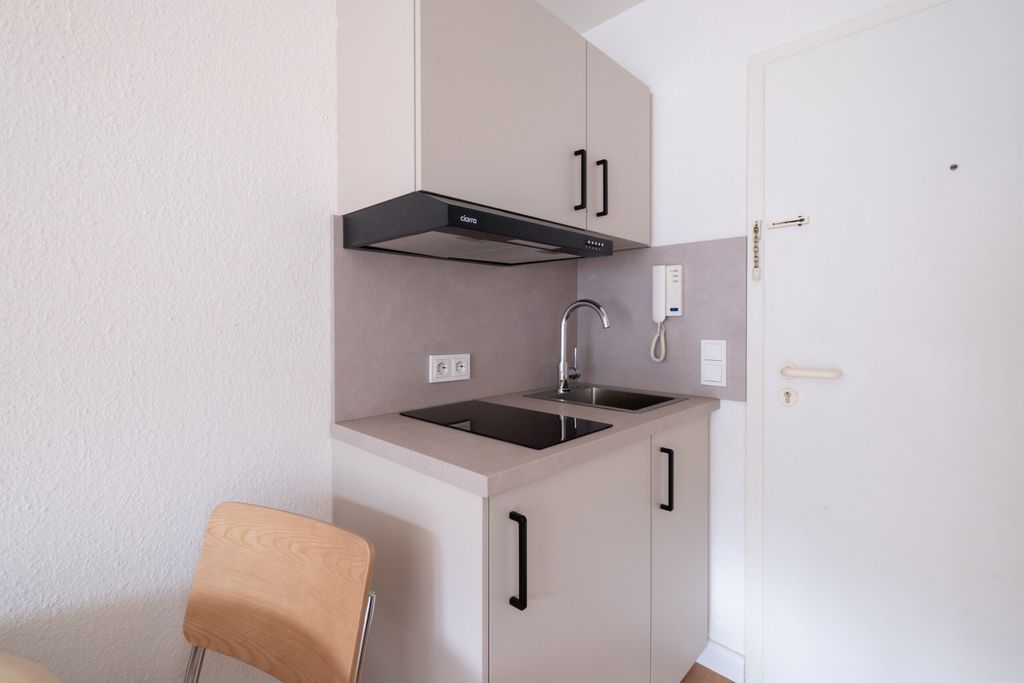 Moderner, kompakter Küchenbereich mit Induktionskochfeld, Spüle, grauer Arbeitsplatte und Hängeschränken, in einem hellen Raum mit weißer Tür.