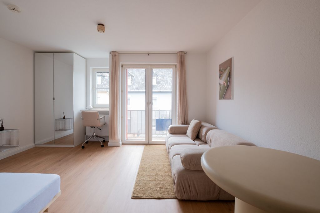 Helles, minimalistisches Wohnzimmer mit beiger Couch, rundem Tisch, Schreibtischstuhl vor Fenster und großem Spiegelkabinett.