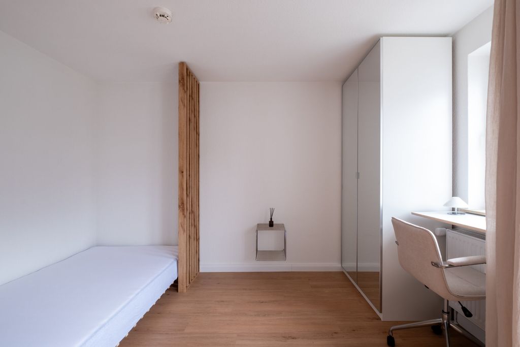 Minimalistisches, kleines Zimmer mit weißem Bett, hölzernem Lamellenelement, eingebautem weißen Schrank mit Spiegel und einem Schreibtisch an einem Fenster mit Stuhl und Lampe.