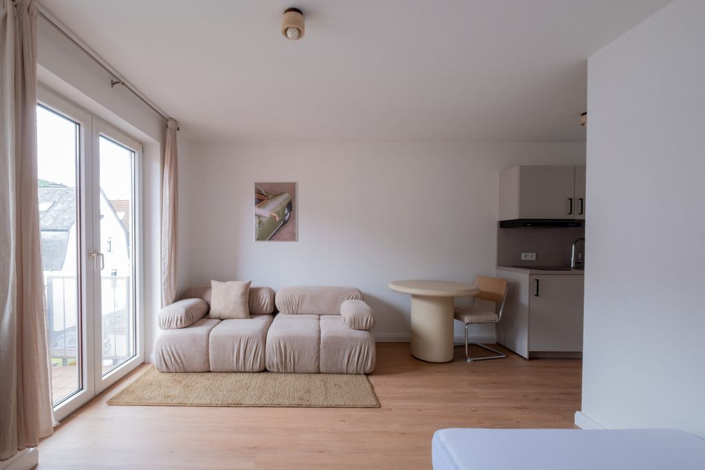 Ein heller Wohnraum mit einem beigen Sofa, einem kleinen runden Esstisch und einem Stuhl neben einer kleinen Küchenzeile, große Fenster mit Vorhängen lassen Tageslicht herein.