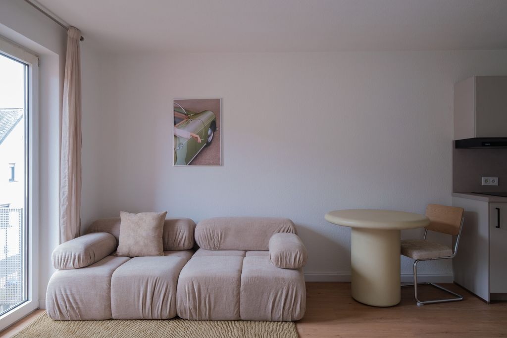 Ein modernes Wohnzimmer mit einem beigen, gemütlichen Sofa, einem runden weißen Tisch und einem Stuhl vor einer weißen Wand mit einem kleinen Bild einer grünen Autotür. Ein großes Fenster lässt natürliches Licht herein.