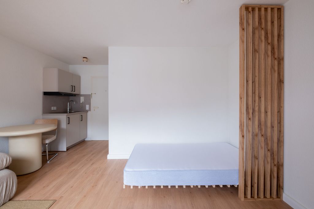 Moderner, minimalistischer Wohnraum mit einem einfachen Bett ohne Bettwäsche, einer kleinen Küchenzeile und einem runden Esstisch mit Stühlen. Der Raum hat helle Holzböden und eine vertikale Holzverkleidung an der Wand neben dem Bett.