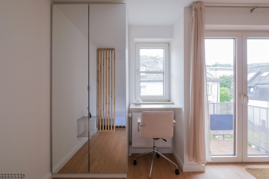 Heller, moderner Arbeitsbereich mit schmalem Schreibtisch, ergonomischem Büro-Stuhl vor einem Fenster und einem Glasschrank mit Spiegel an der Wand.