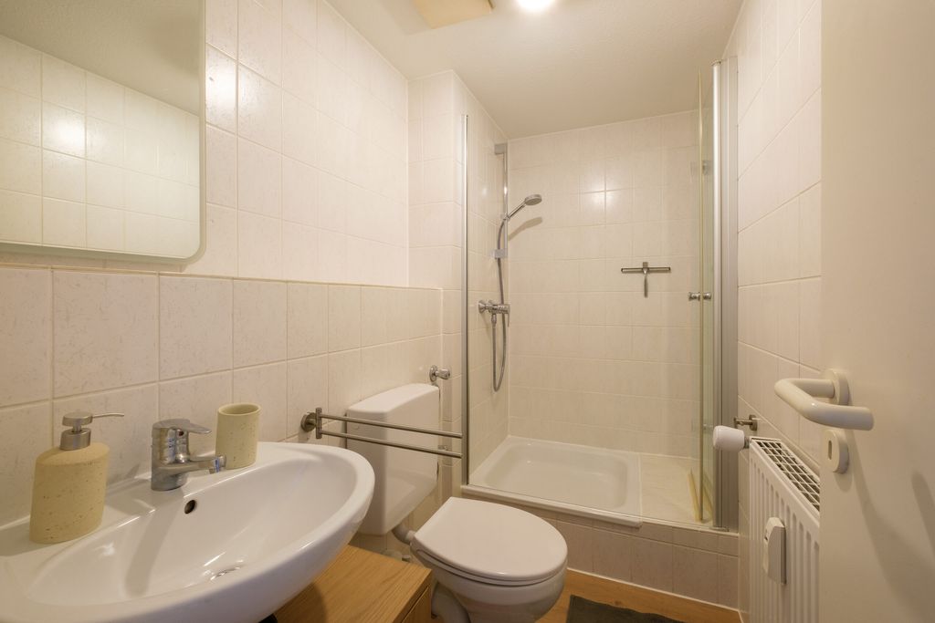 Kleines Badezimmer mit weißer Toilette, Waschbecken, beigem Seifenspender und Duschkabine mit Glasrahmen.