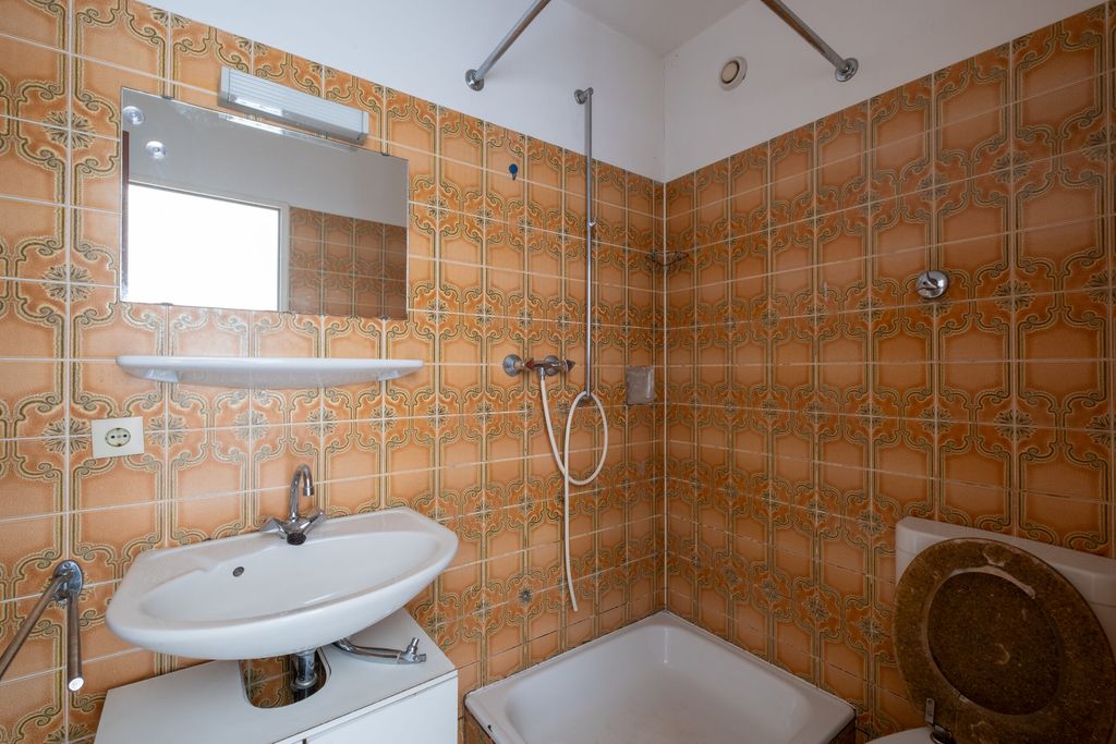Badezimmer mit orangefarbenen Fliesen und Mustern, Dusche mit Handbrause, weißem Waschbecken und Toilettensitz mit Holzdeckel.