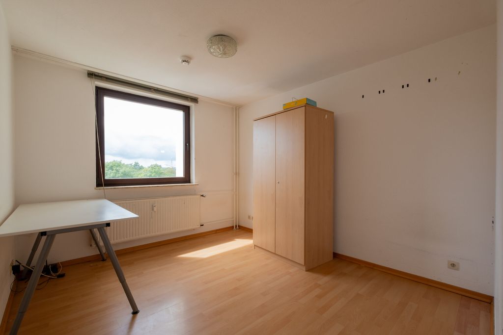 Leerer Raum mit hellen Holzböden, einem weißen Tisch mit grauen Beinen und einem Holzkleiderschrank vor einer weißen Wand mit einem Fenster, das Tageslicht hereinlässt.