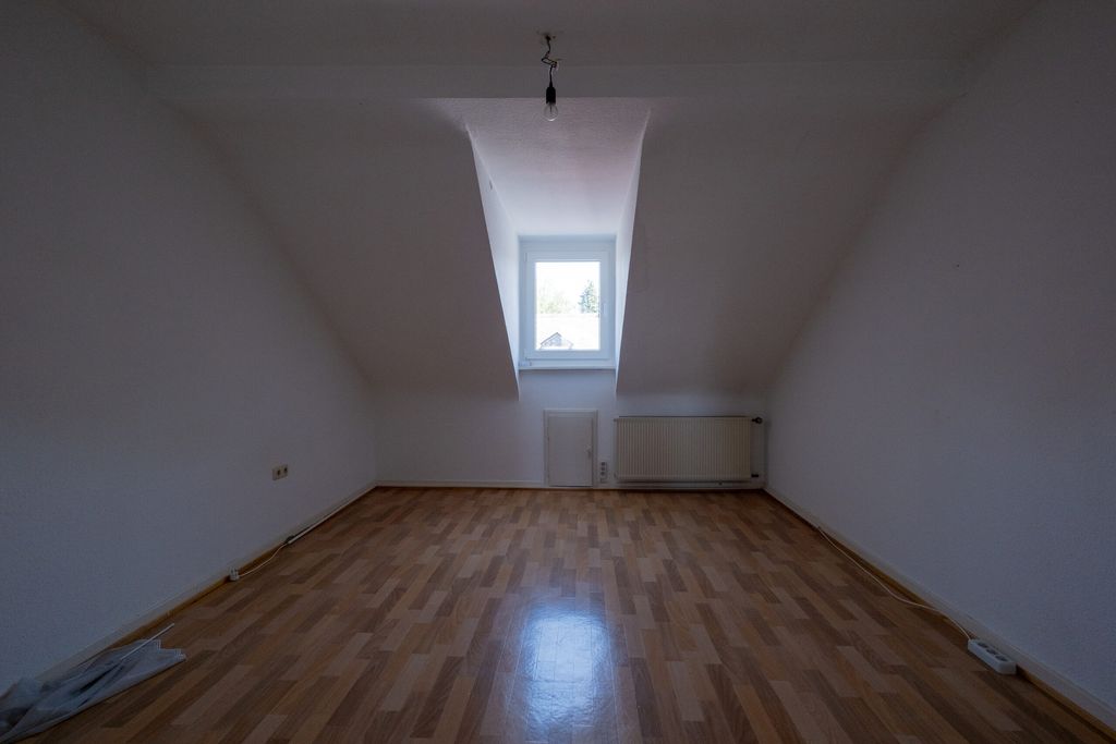 Leerer Raum mit hellbraunem Laminatboden, weißen Wänden, Dachschräge und einem Fenster mit Blick nach draußen.