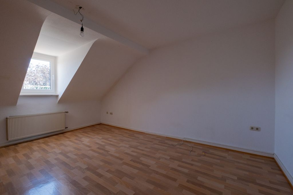 Leerer Raum mit hellbraunem Laminatboden, weißen Wänden, einem kleinen Fenster unter einer Dachschräge sowie einem Heizkörper unter dem Fenster.