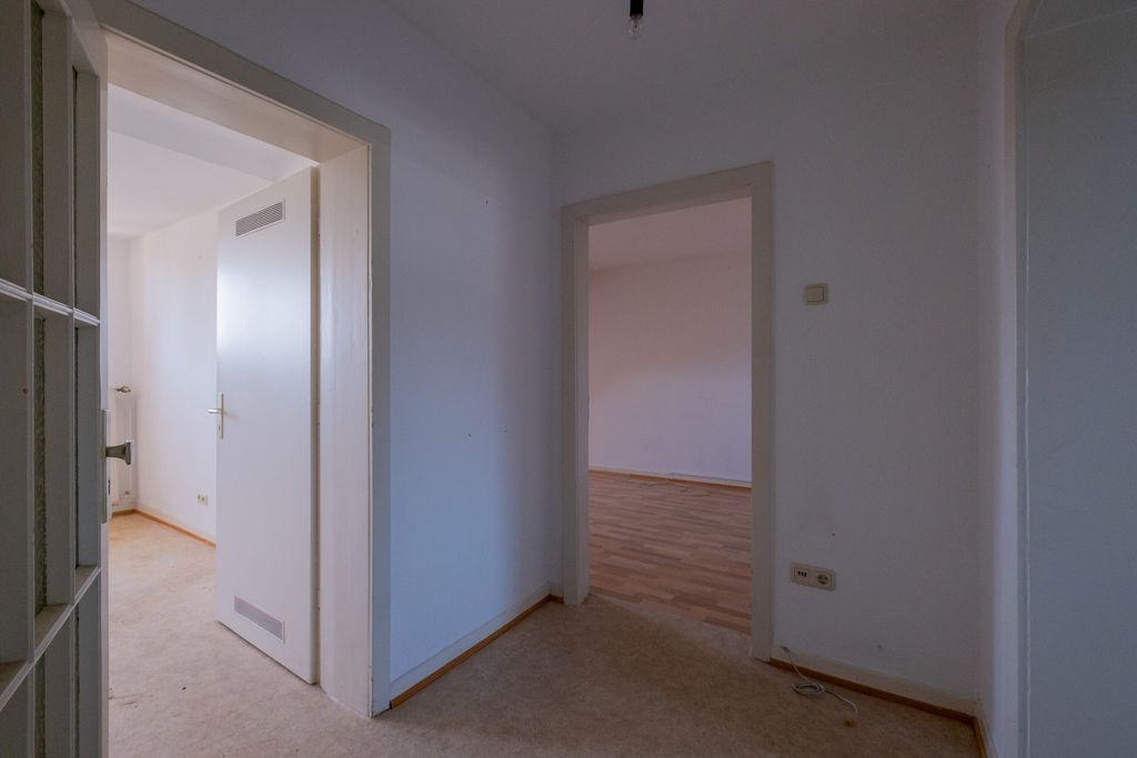 Blick in einen leeren Flur mit zwei offenen Türen, einer führt in einen Raum mit Laminatboden, der andere in einen Raum mit hellem Teppichboden.