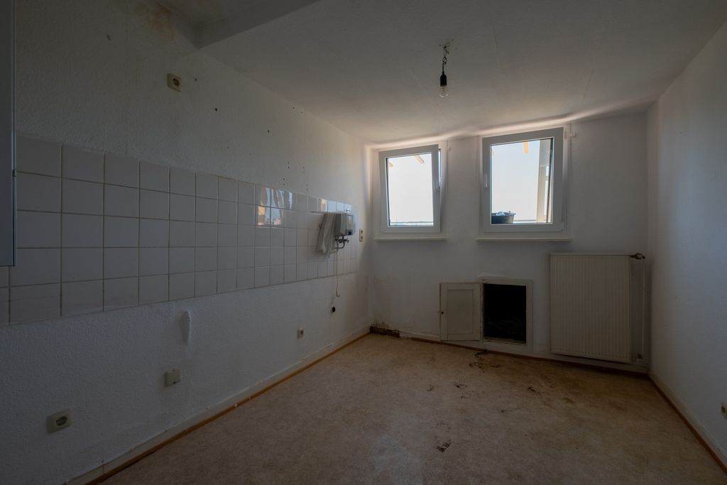 Leerer, dunkler Raum mit weißen Fliesen an einer Wand, zwei kleinen Fenstern und Heizkörper an der Wand.