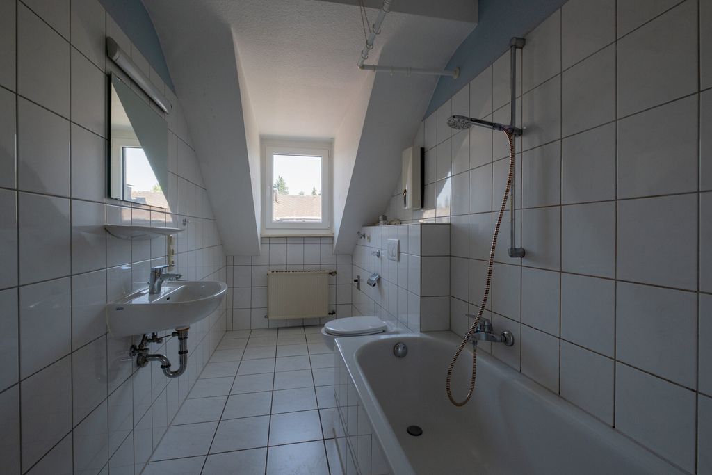 Kleines Badezimmer mit weißen Fliesen, einer Badewanne mit Duschkopf, einem Waschbecken, einer Toilette und einem Fenster unter einer Dachschräge.