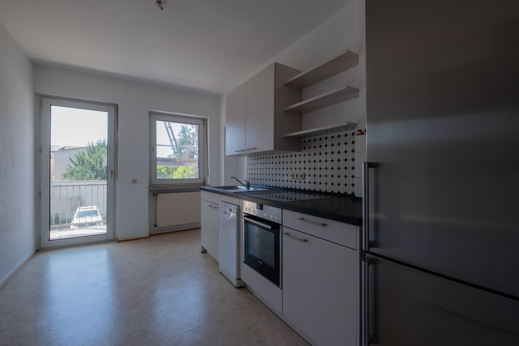 Moderne, helle Küche mit weißen Schränken, eingebautem Backofen, Geschirrspüler, großem Fenster und Zugang zu einem Balkon mit Blick auf eine Straße und Nachbargebäude.