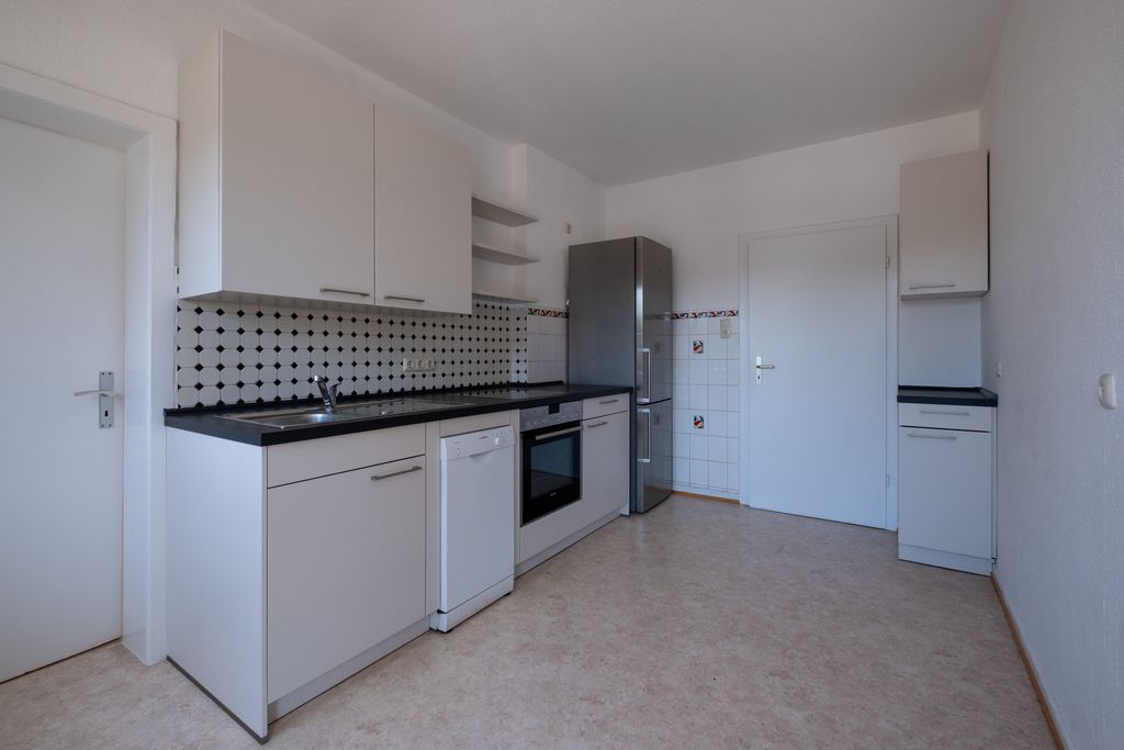 Moderne weiße Küche mit schwarzen Arbeitsplatten, eingebautem Ofen, Geschirrspüler und großem silbernen Kühlschrank in einem hellen Raum mit weißen Wänden und Tür.