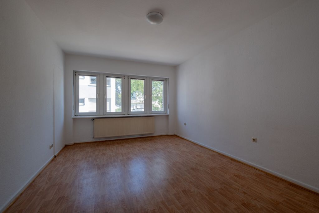 Leerer Raum mit Holzfußboden, weißen Wänden, großen Fenstern und einem Heizkörper unter den Fenstern.