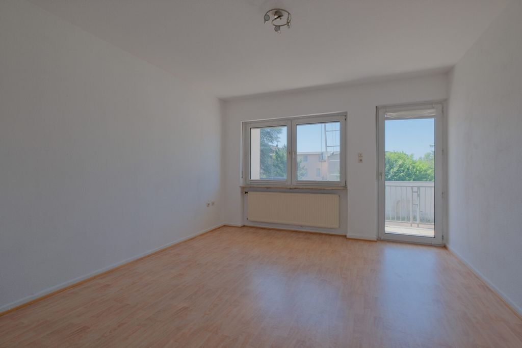 Leerer Raum mit hellen Holzboden, weißen Wänden, einem Fenster und einer Tür, die zu einem kleinen Balkon führt.