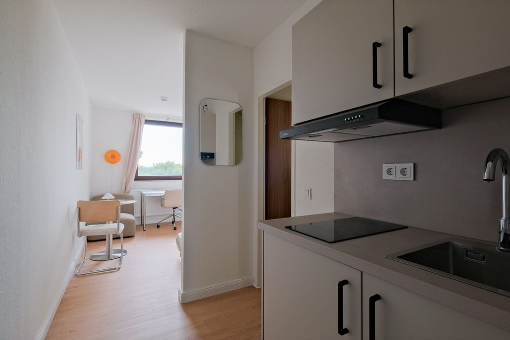 Kleine helle Küchenzeile mit elektrischer Kochplatte und Spüle, angrenzend ein Wohnbereich mit Stühlen, Tisch und Fenster mit Vorhängen.