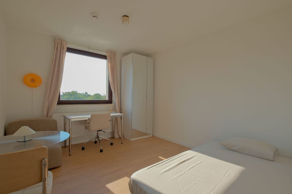 Hell eingerichtetes Zimmer mit großem Fenster, Schreibtisch mit Stuhl, Kleiderschrank mit Spiegeltür, gemütlichem Sessel und einem Bett mit einem Kissen.