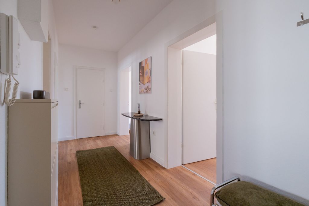 Flur mit Holzboden, einem grünen Teppich, einem kleinen Tisch mit Dekorationsgegenstand und weißen Türen in einer hellen Wohnung.