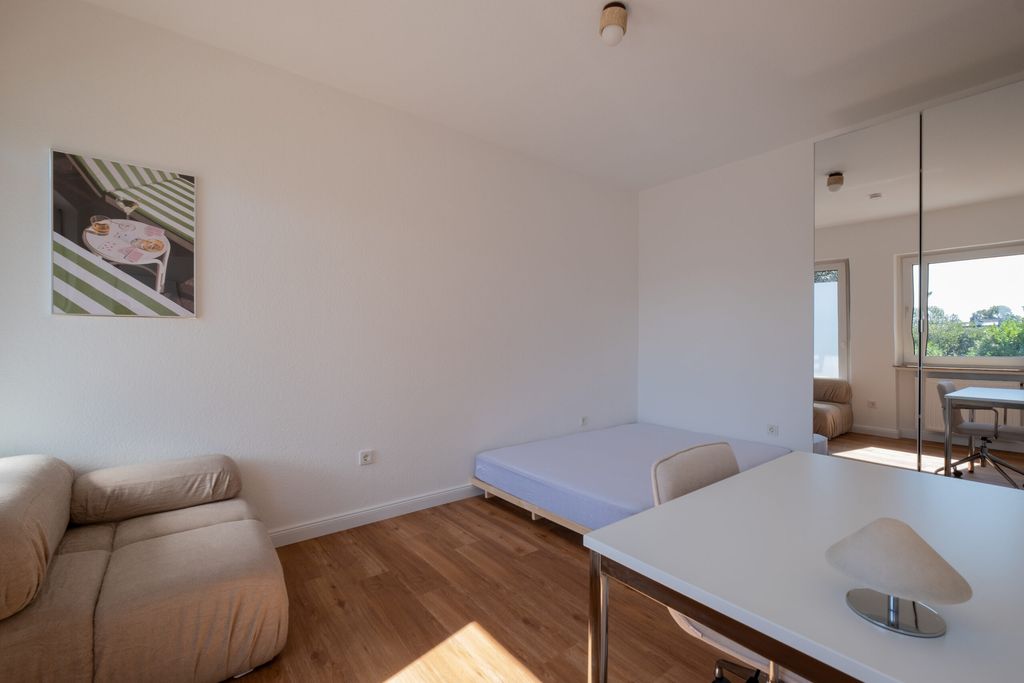 Ein einfach eingerichtetes Zimmer mit einem grauen Sofa, einem niedrigen Bett ohne Bettwäsche, einem weißen Tisch mit Stuhl und einem Spiegelkleiderschrank, der das Fenster und die Außenansicht reflektiert.