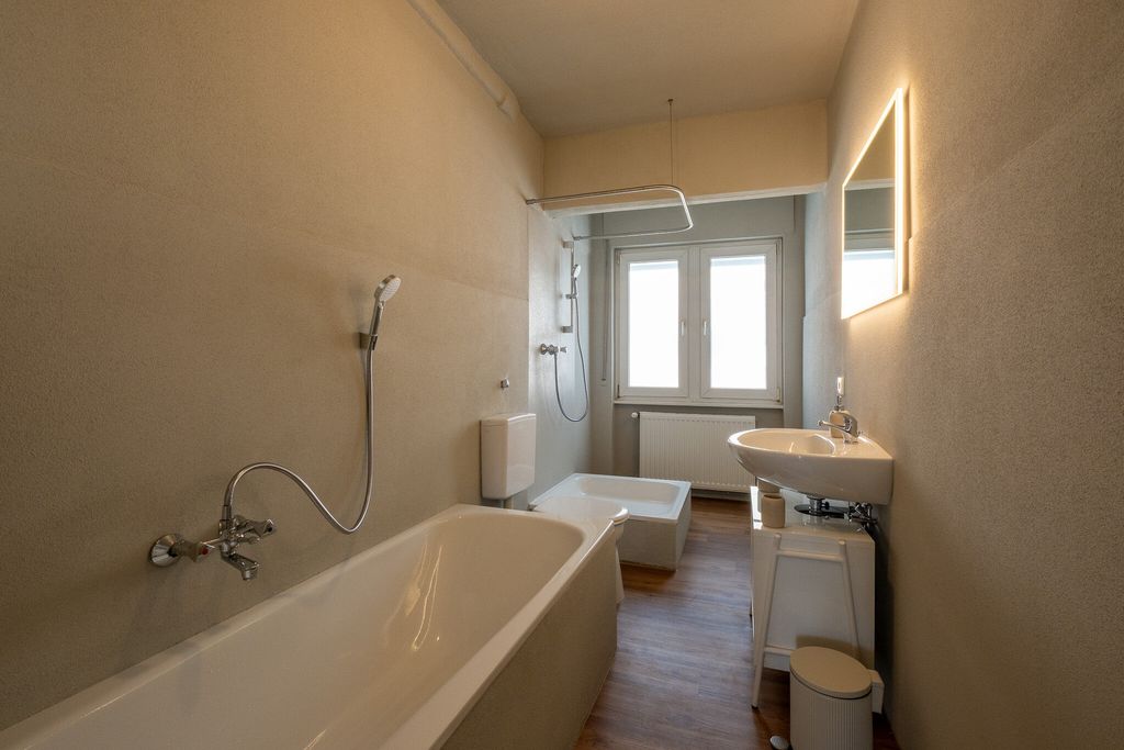 Ein schmales Badezimmer mit Badewanne, Waschbecken, beleuchtetem Spiegel und einer Dusche vor einem Fenster mit natürlichem Licht.