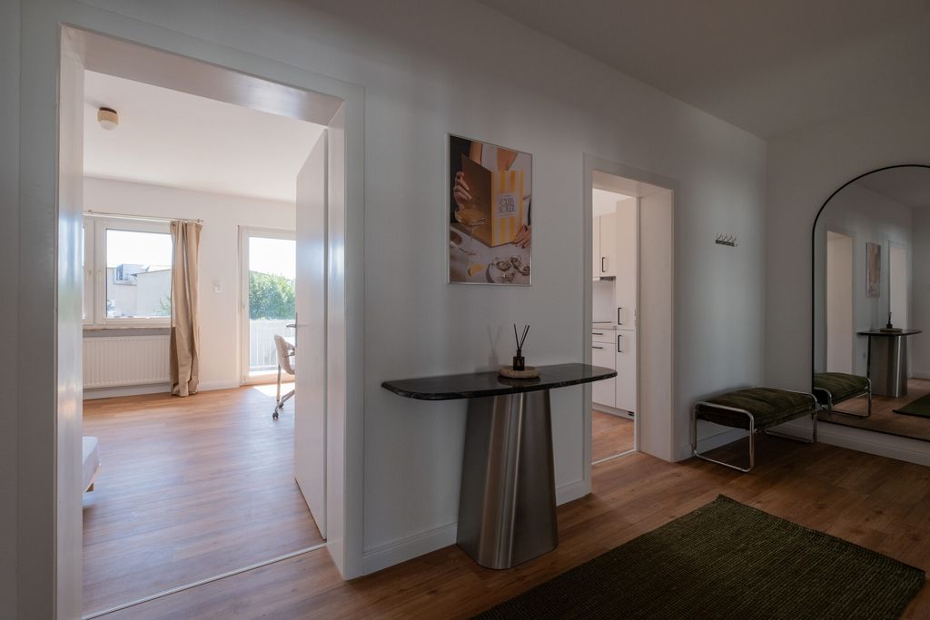 Moderner Flur mit hellem Holzboden, einem schwarzen Konsolentisch, einer Fotografie an der Wand, einem grünen Teppich und einer großen Wandspiegel, der einen grünen Sitzhocker reflektiert. Blick in ein helles Zimmer mit Stuhl am Fenster und eine weiße Küche.