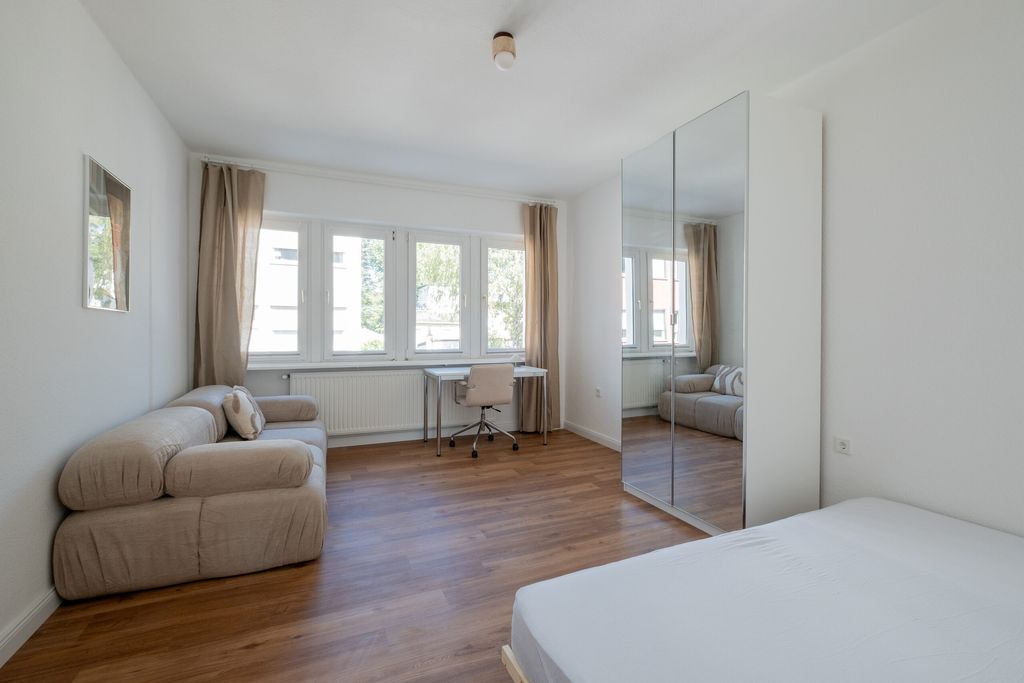 Helles Schlafzimmer mit Holzboden, Doppelbett, beigem Sofa, Schreibtisch vor dem Fenster und einem Kleiderschrank mit Spiegeltüren.