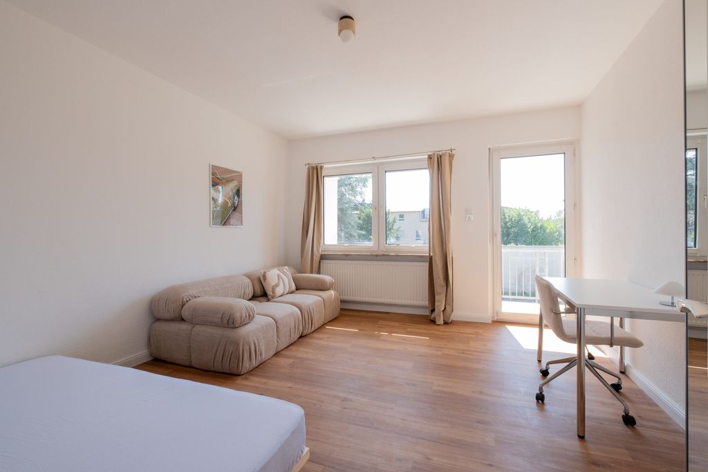 Helles, modernes Wohnzimmer mit einem beigen Sofa, einem Schreibtisch mit Stuhl, einem Fenster mit Vorhängen und Holzboden.