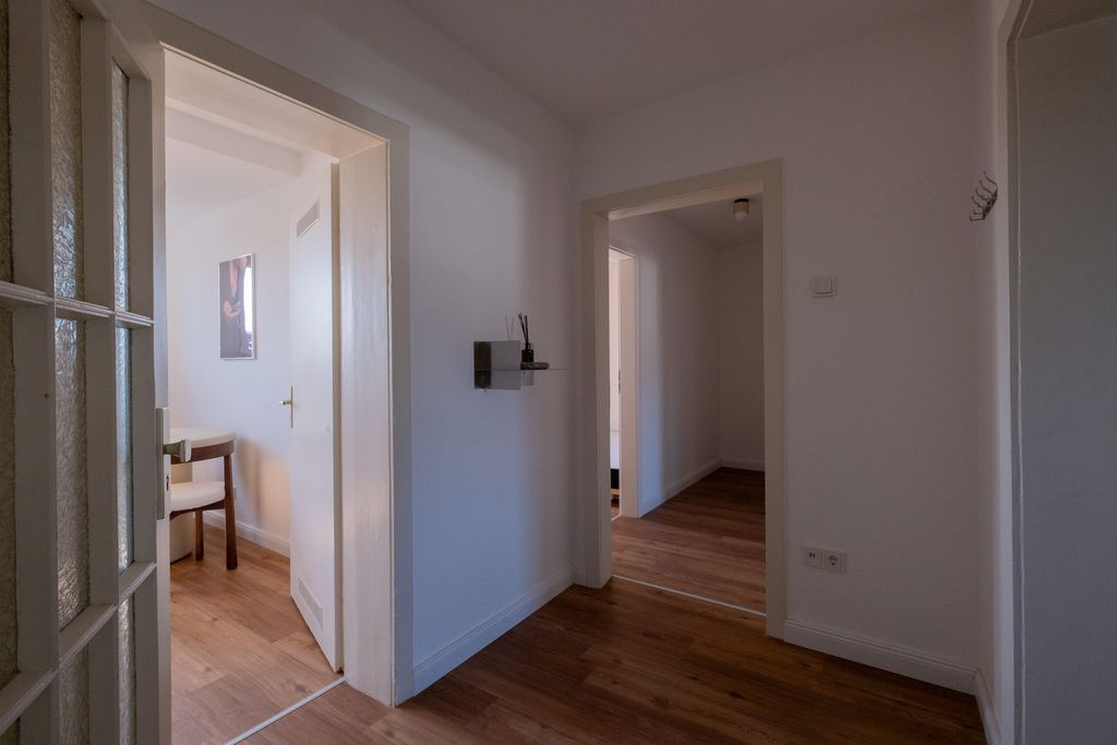 Flur mit Holzboden und weißen Wänden, der zu zwei angrenzenden Räumen mit Holzboden und weißen Wänden führt, einer mit einem Stuhl und einem Bild an der Wand.