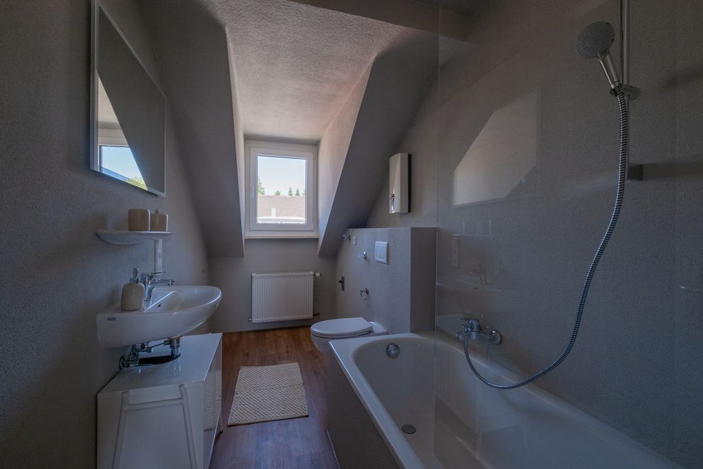 Kleines Badezimmer unter einer Dachschräge mit Badewanne, Dusche, Waschbecken und Toilette, ein kleines Fenster ist im Hintergrund sichtbar.
